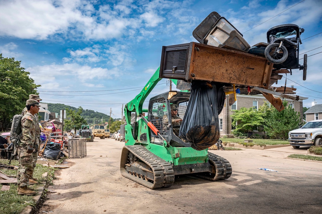 A man in a camouflage military uniform removes furniture and other debris with a waste handler track loader on a neighborhood street, as two other men in similar attire observe from the curb.
