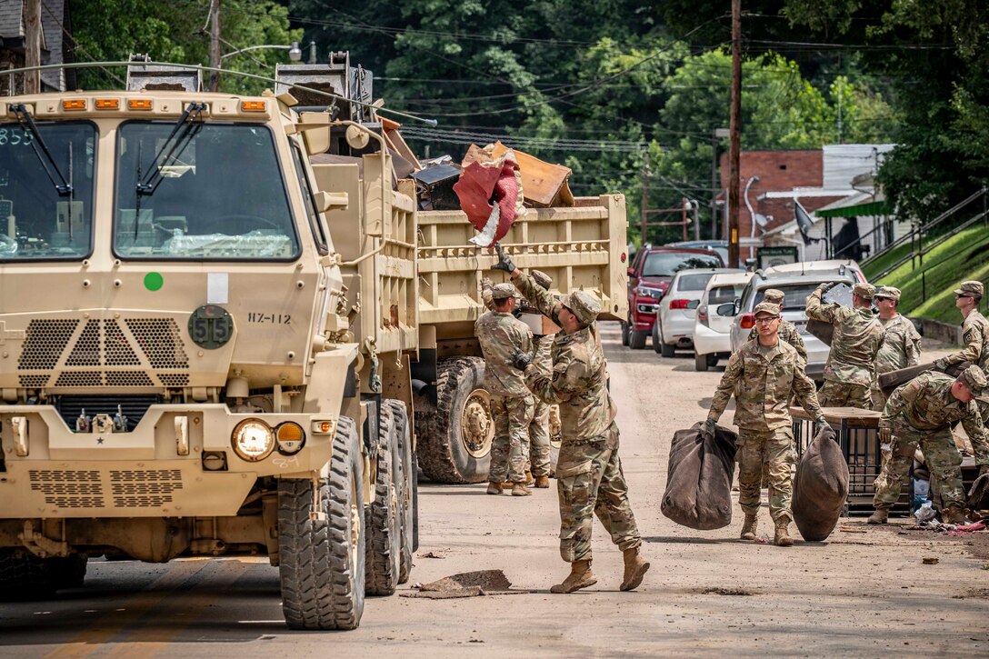 Nearly a dozen people in camouflage military uniforms pick up debris from the side of a street and throw it into the backs of two large military dump trucks.