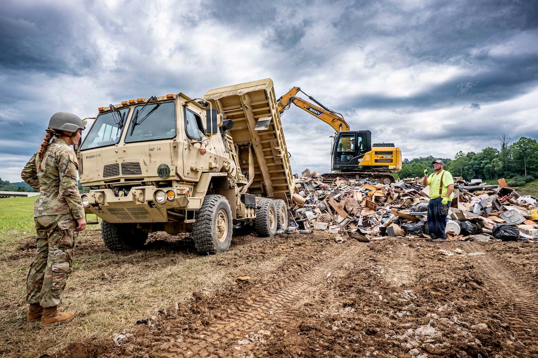 A woman in a camouflage military uniform stands in front of a military dump truck that has dumped a large pile of debris onto a field as a man in casual attire stands to the right of the truck signaling the driver. Construction equipment and a marbled dark blue and white sky are in the background.