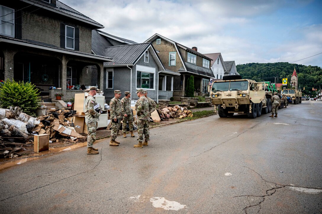 Five people in camouflage military uniforms talk on a cracked mud-covered street lined with homes.