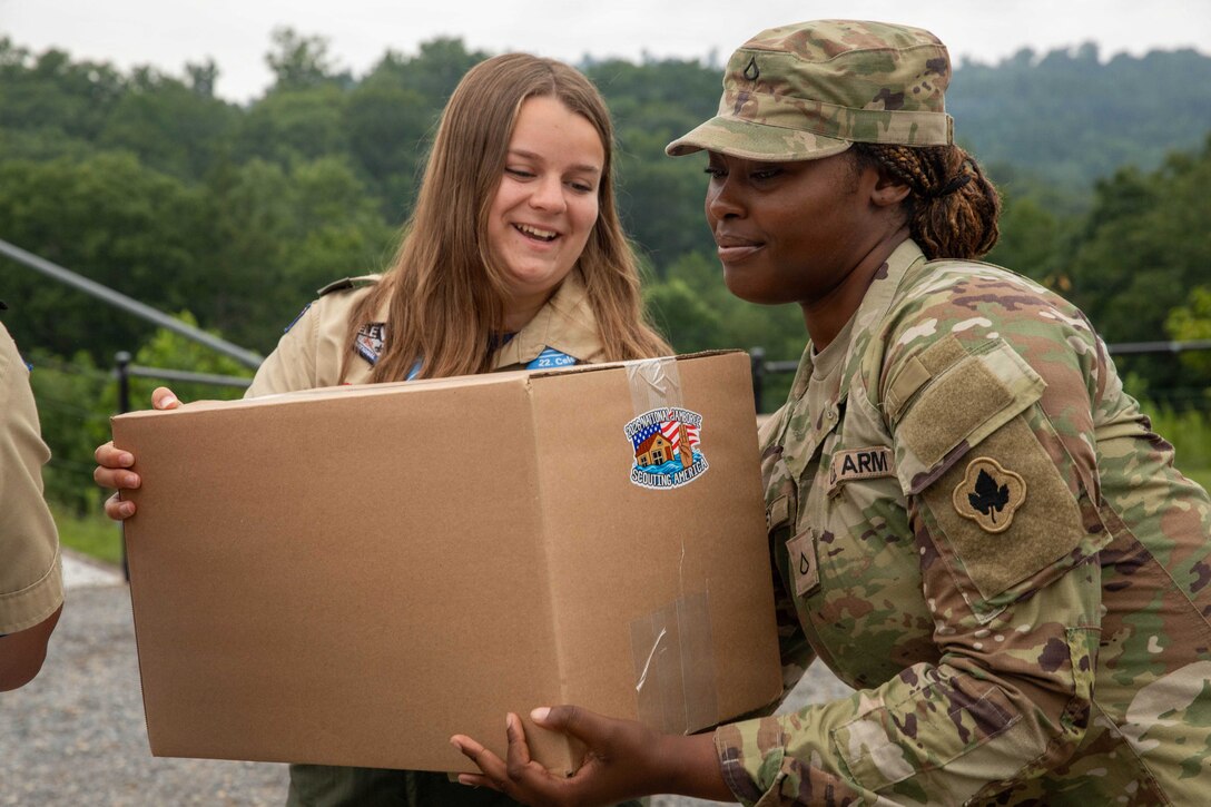 A woman in a camouflage military uniform hands a cardboard box to a young girl outside; trees are in the background.