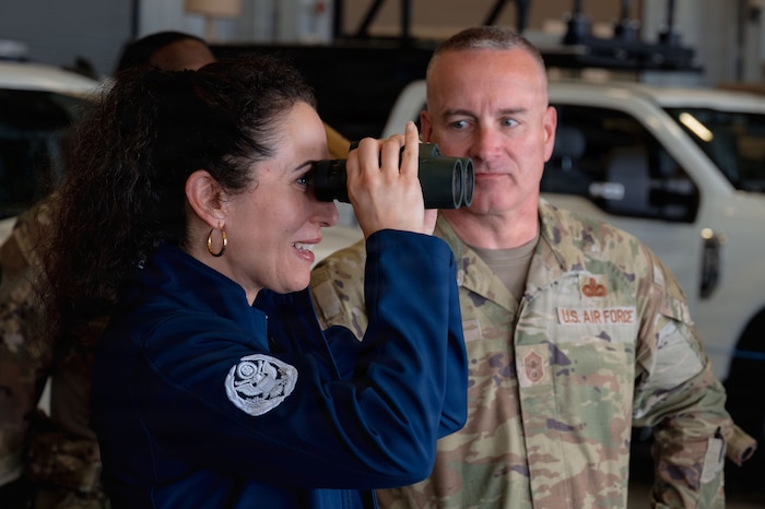A photo of Dr. Doniel Wolfe looking through binoculars while being watched by Chief Master Sergeant of the Air Force David R. Wolfe.