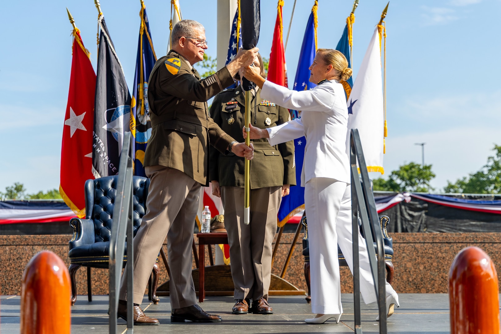 A man in a Army uniform hands a flag to a woman in a white Navy uniform.