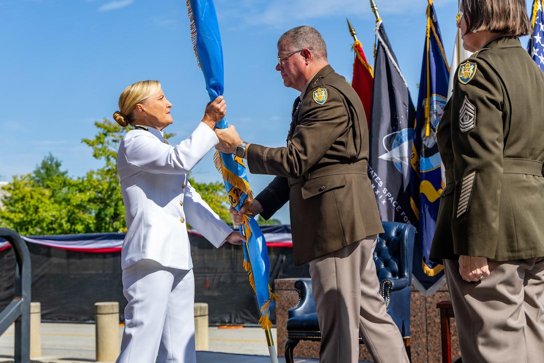 A man in a Army uniform hands a flag to a woman in a white Navy uniform.