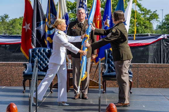 A woman in a Navy uniform passes a blue flag to a woman in a Army uniform before a man in a Army uniform on a dark stage with a fan of flags behind them. The stage is outside underneath a large flag pole. 