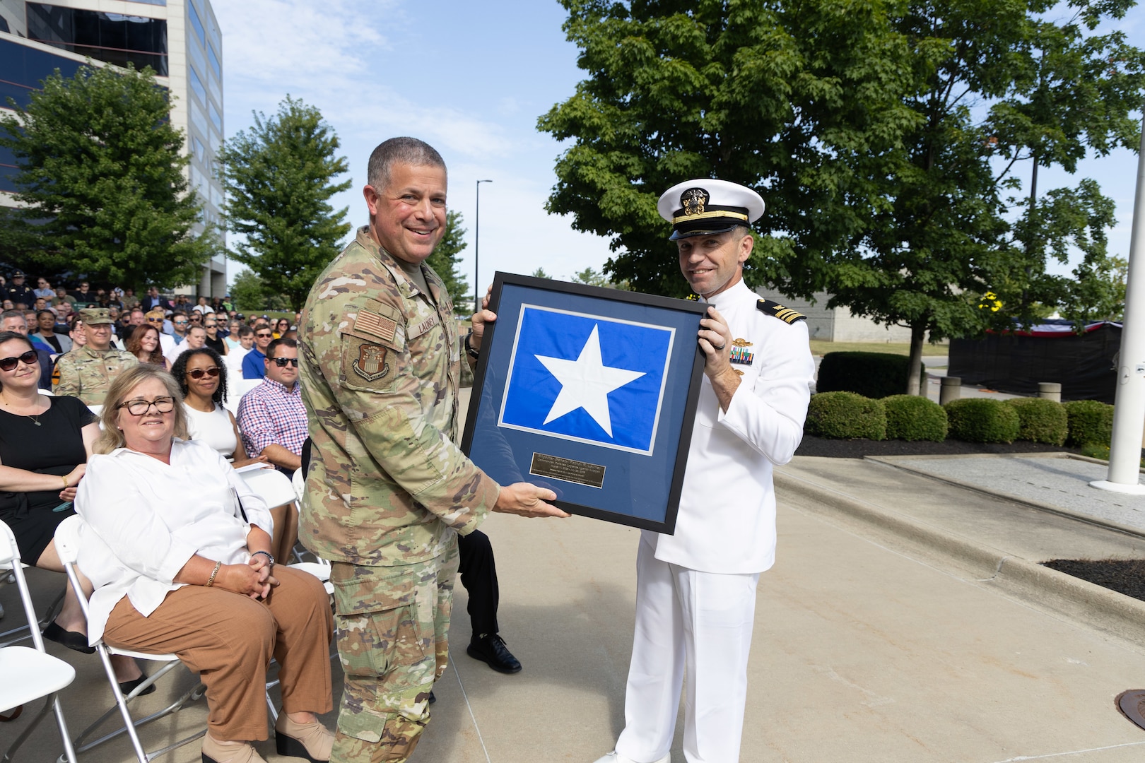 A man in a Air Force utility uniform holds a framed flag with a man in a white Navy uniform.