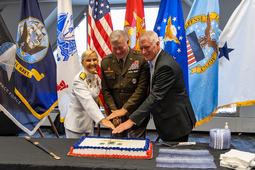 A woman in a white dress Navy uniform, a man in a dress Army uniform and a man in a dark suit cut a cake with a sword.
