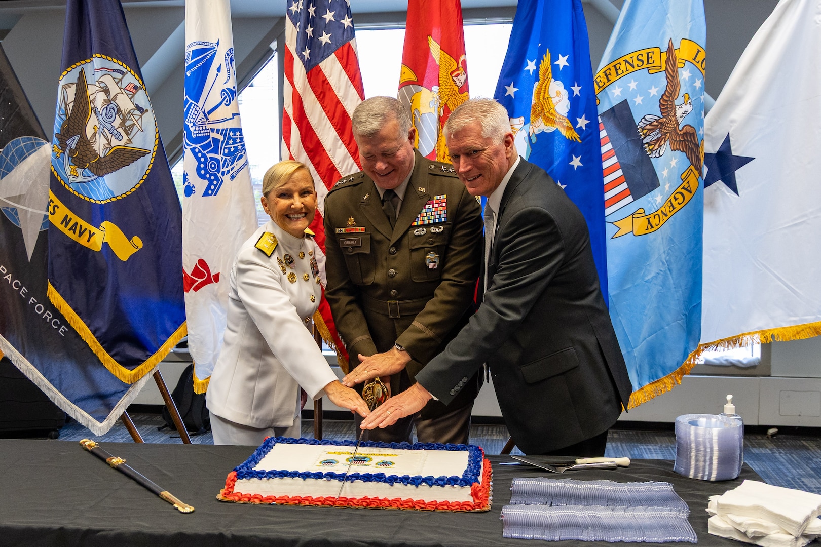 A woman in a white dress Navy uniform, a man in a dress Army uniform and a man in a dark suit cut a cake with a sword.