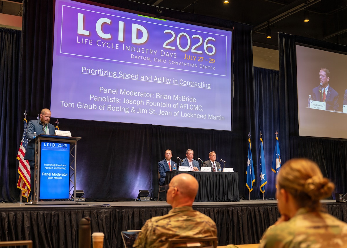 A stage during a panel event. Uniformed service members watch from the audience.