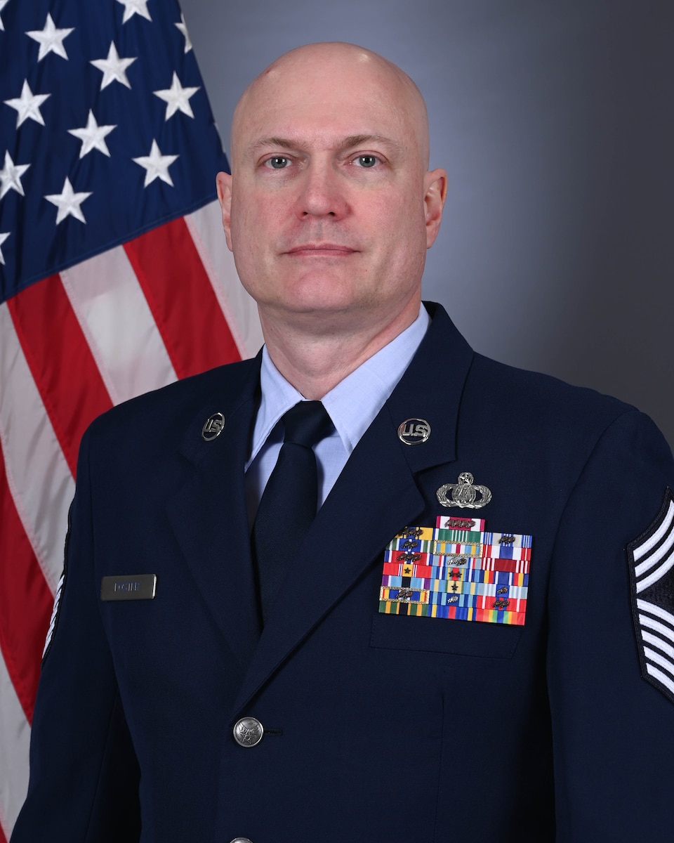 Official Photo of CMSgt Jason Foster. He is wearing service blues and standing in front of an American Flag.
