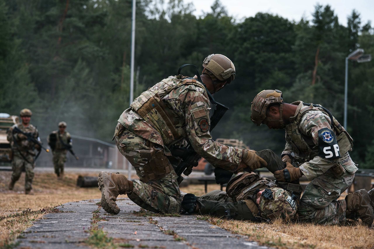 Two people in camouflage military uniforms check on a person in similar attire who is lying on the ground.