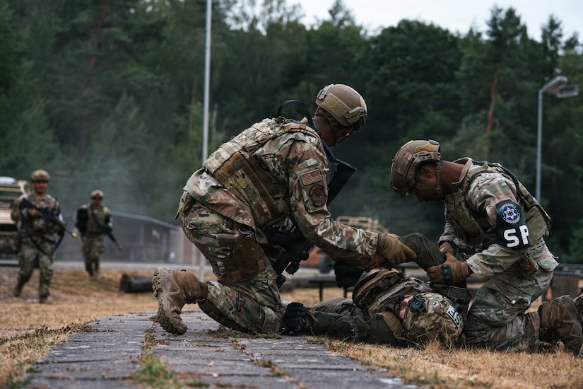 Two people in camouflage military uniforms check on a person in similar attire who is lying on the ground.