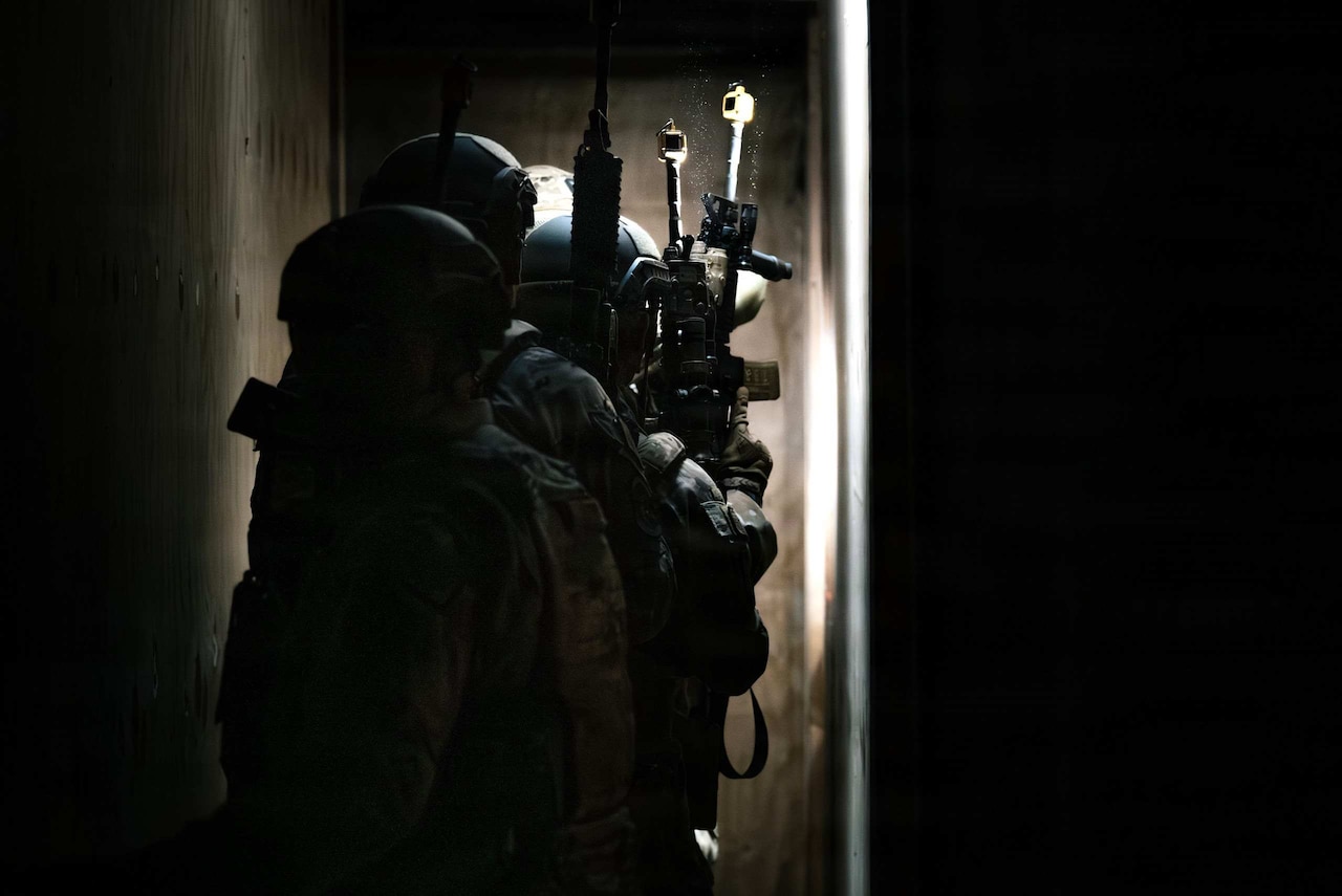 Four people in camouflage military uniforms hold rifles and walk down a dark hallway.