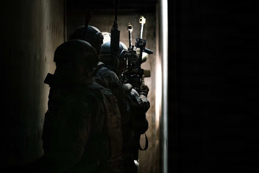 Four people in camouflage military uniforms hold rifles and walk down a dark hallway.