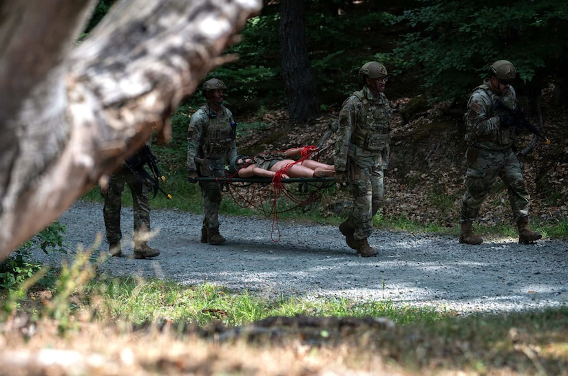 Two men in camouflage military uniforms carry a mannequin on a stretcher while walking in the woods; two other people in similar attire hold rifles and walk beside them.