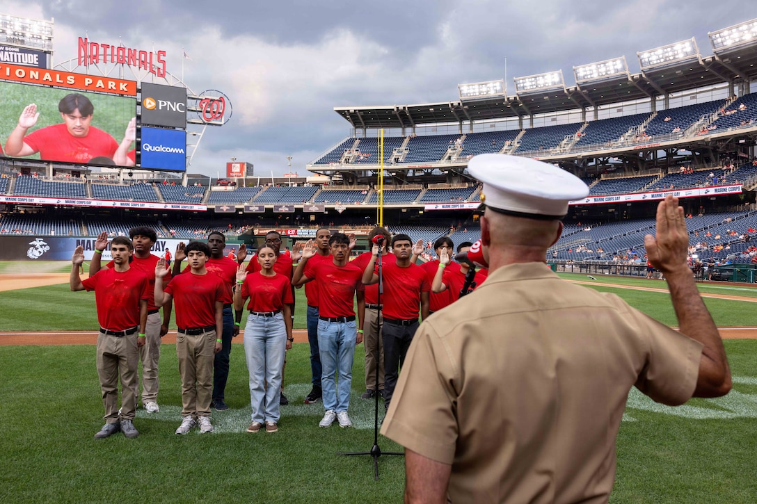 A Marine raises their right hand while facing a dozen poolees in red shirts raising their right hands on a baseball field with an empty stadium.