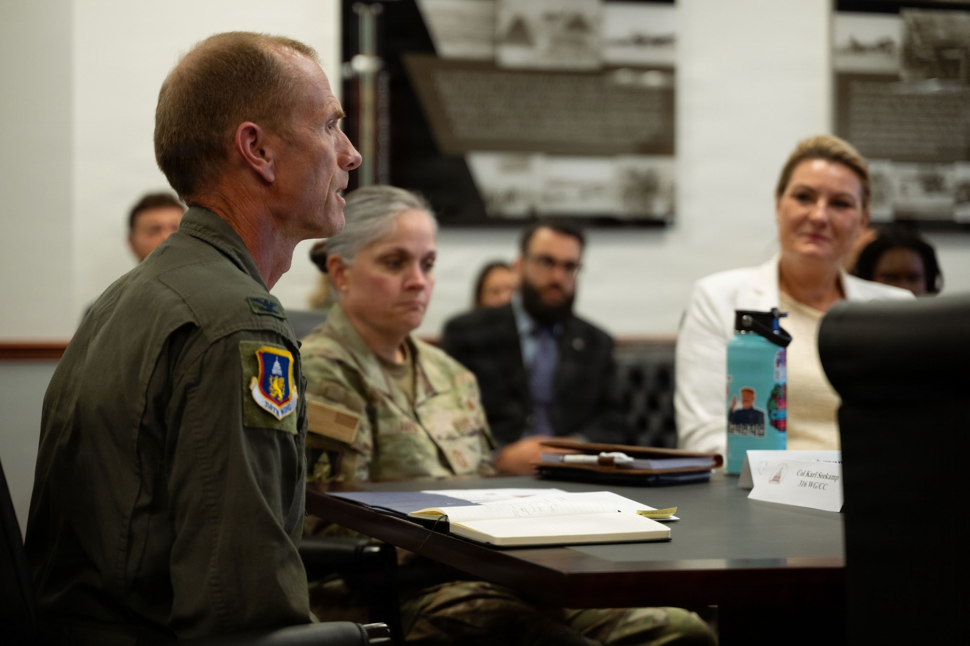 A man in a green flight suit sits at the head of a table and speaks to a group of people also seated.
