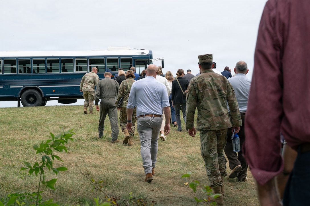 A group of people, some in military garb and others in civilian clothes walk up the hill to a blue bus.