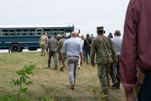 A group of people, some in military garb and others in civilian clothes walk up the hill to a blue bus.