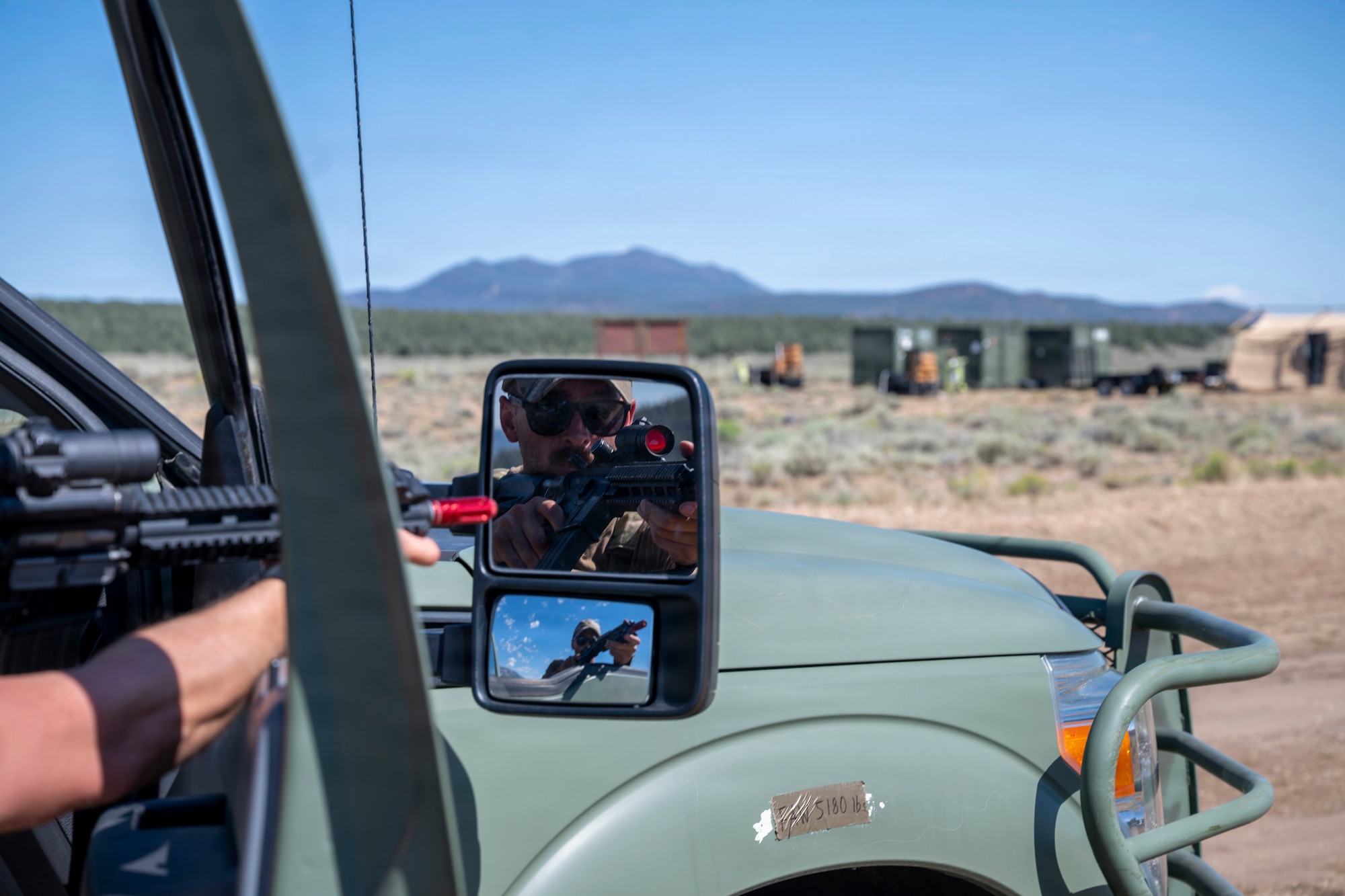 A mirror on a truck displays an airmen holding a weapon during training outside.