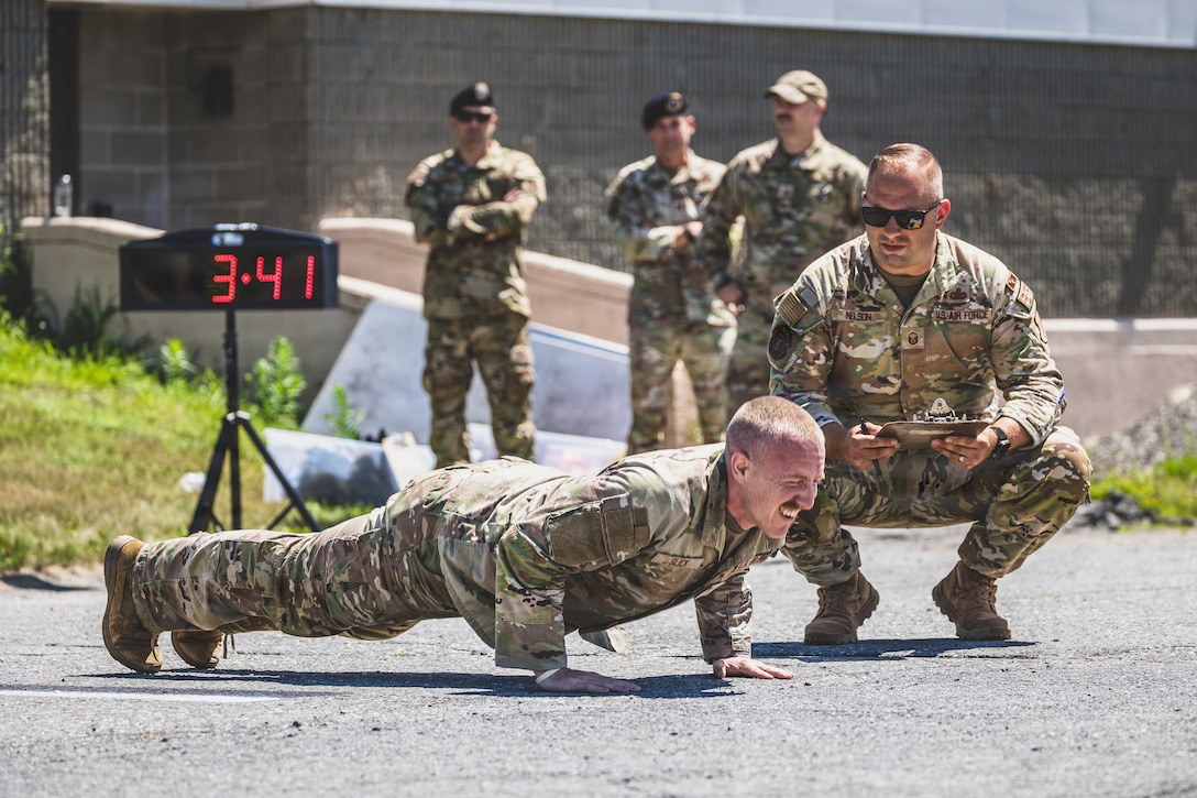 A kneeling airman with a clipboard evaluates an airman as they perform pushups with a digital clock to left and others watching on the right.