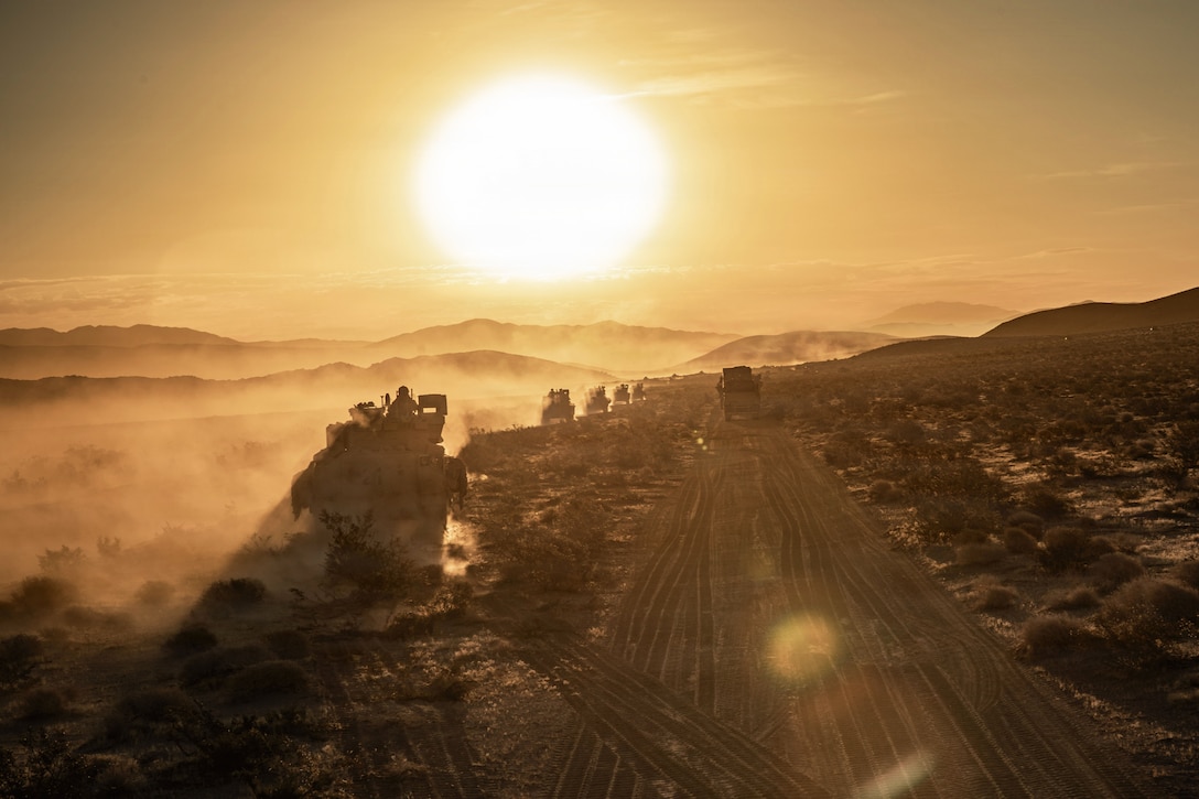 Army tactical vehicles drive through a desert-like area as the golden sun shines over hilly terrain.
