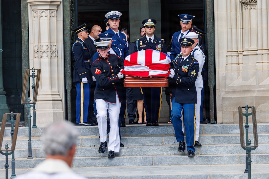 Seven people in military uniforms carry a casket with an American flag over it down steps in front of a church.