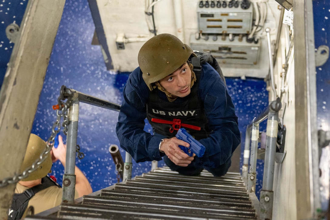 A person in a blue uniform that says U.S. Navy stands on a metal ladder in a ship and looks up while holding a plastic royal blue gun. Another soldier is standing on the floor in the background.