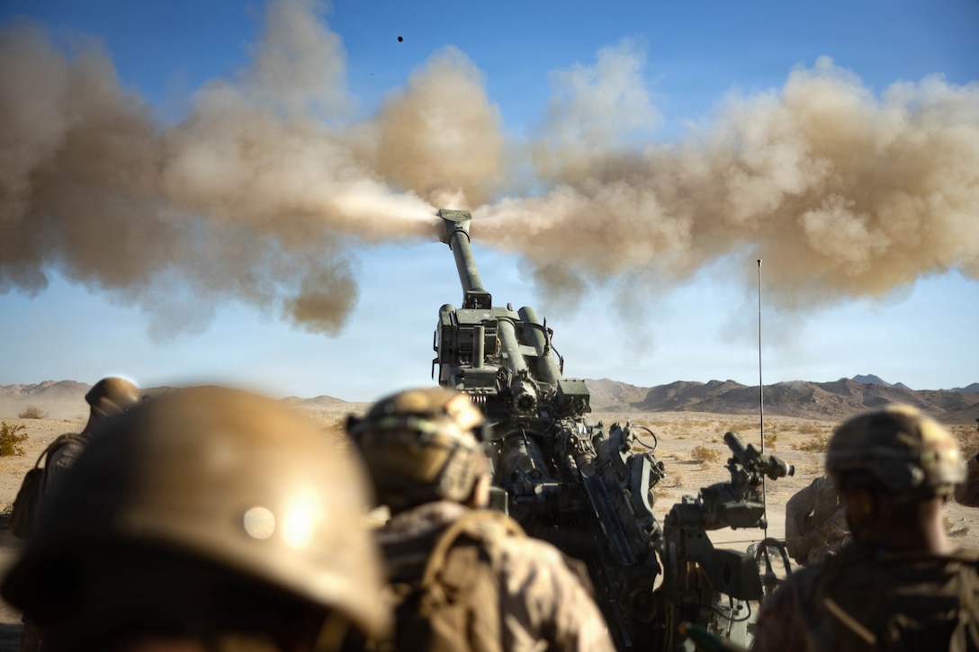 Smoke comes out of a howitzer in the desert as people, in rearview, in camouflage uniforms and helmets stand behind it.