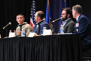 Four men sit at table. Two are wearing Air Force uniforms, the other two business suits. There is a black cloth around the table.