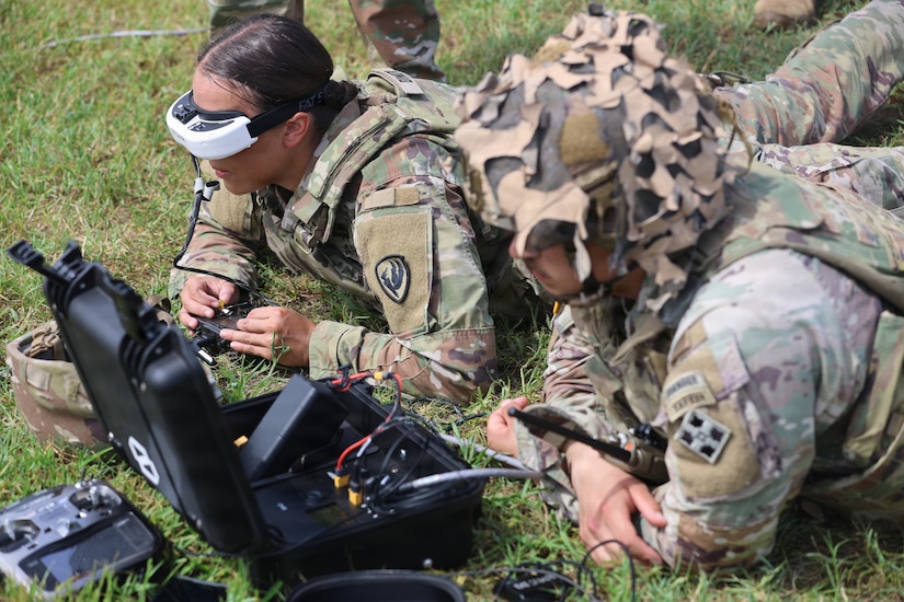A woman wearing a camouflage military uniform and a tactical headset is lying in the grass in the prone position while a man in similar attire and a helmet looks at a computer screen.