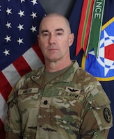 Army soldier wearing Army combat uniform posing for an official photo in front of the U.S. and organizational flags.