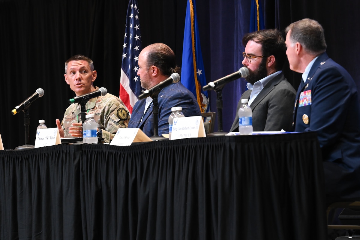 Four men sit at table. Two are wearing Air Force uniforms, the other two business suits. There is a black cloth around the table.