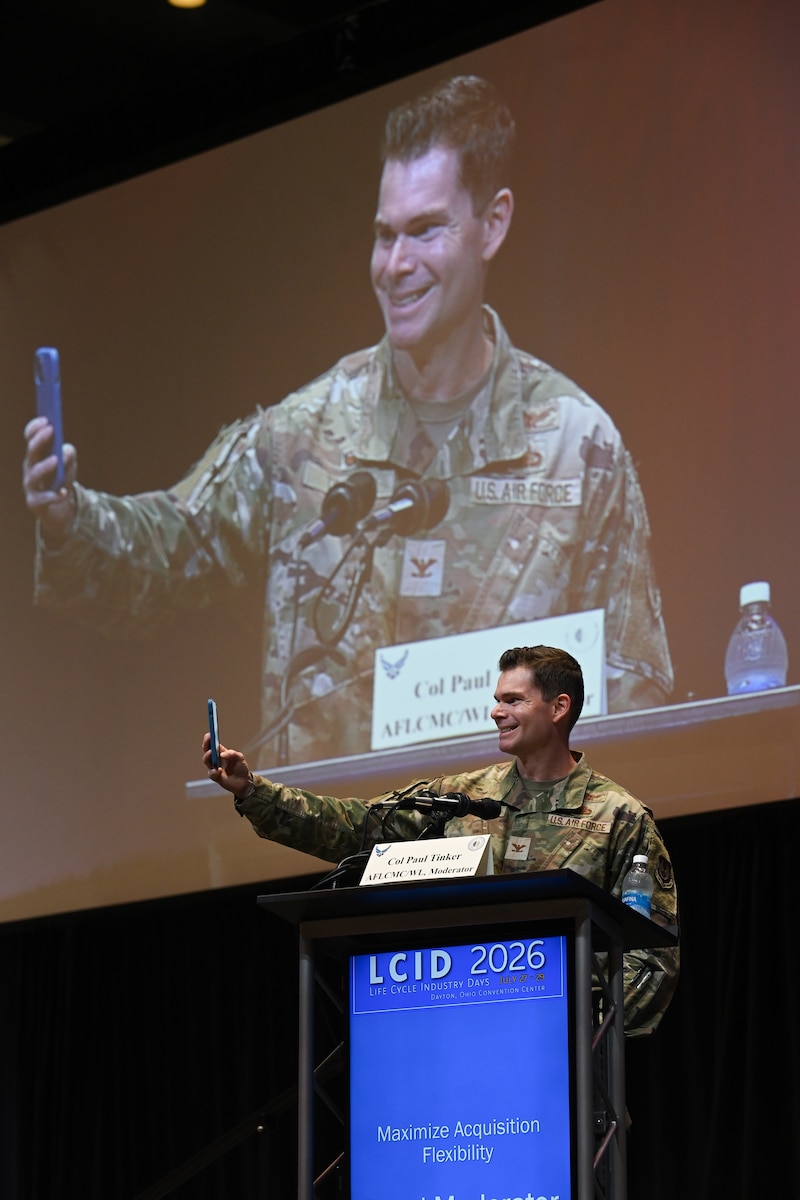 Male colonel in Air Force camo takes selfie on stage, behind podium that reads "LCID"