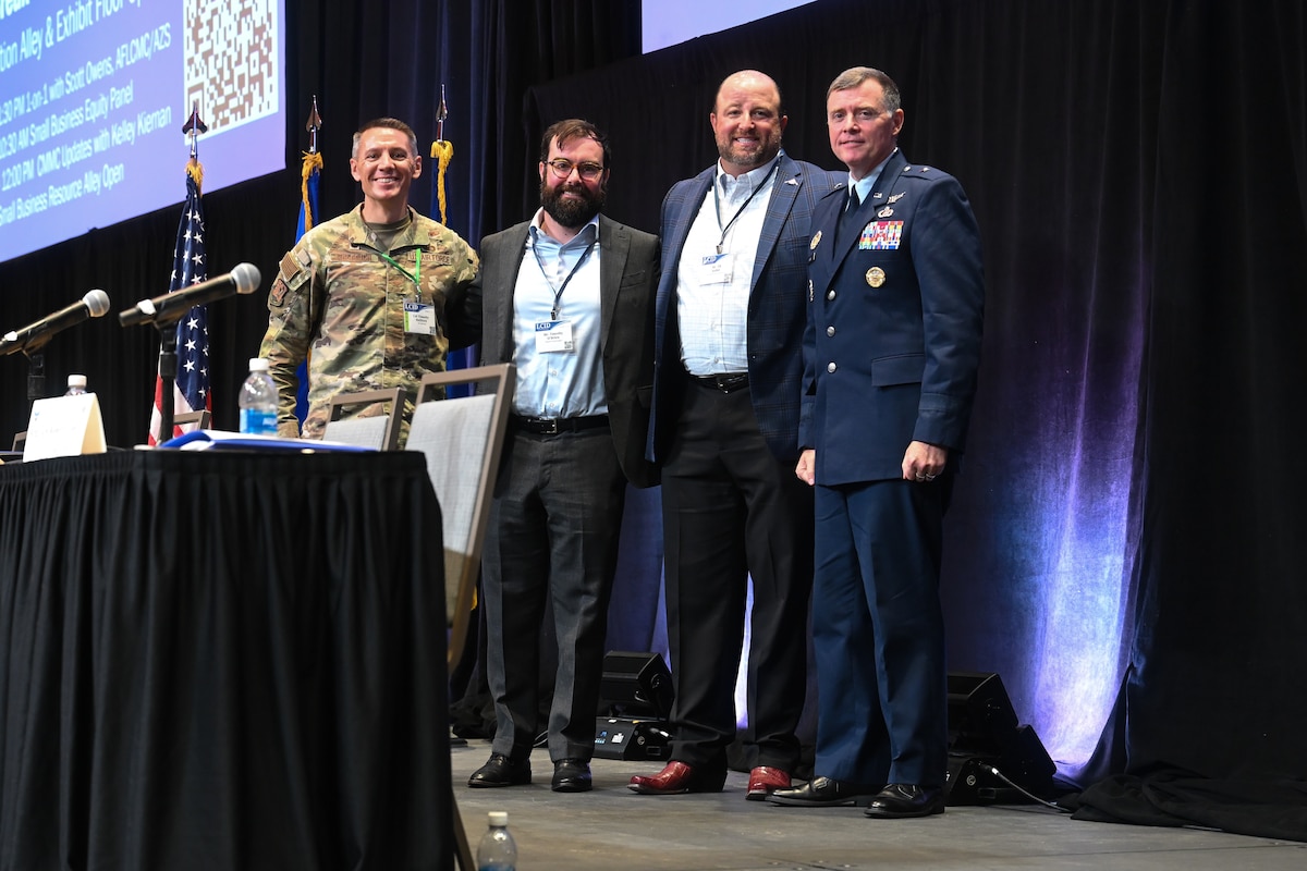Four men stand on stage in front of large screen and pose for photo.
