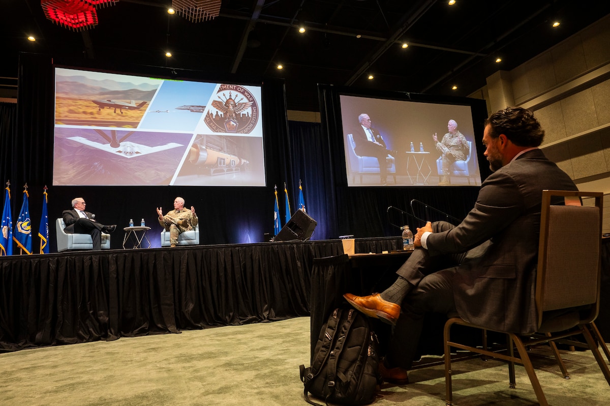 Men sitting on stage, talking in front of large screens showing Air Force planes.