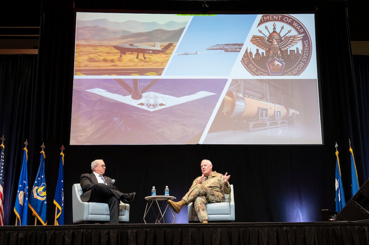 Men sitting on stage, talking in front of large screens showing Air Force planes.