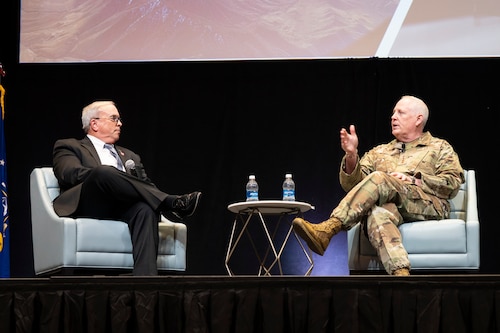 Men sitting on stage, talking in front of large screens showing Air Force planes.