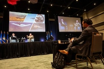Men sitting on stage, talking in front of large screens showing Air Force planes.