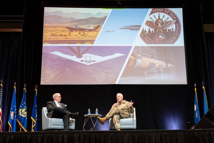 Men sitting on stage, talking in front of large screens showing Air Force planes.