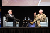 Men sitting on stage, talking in front of large screens showing Air Force planes.