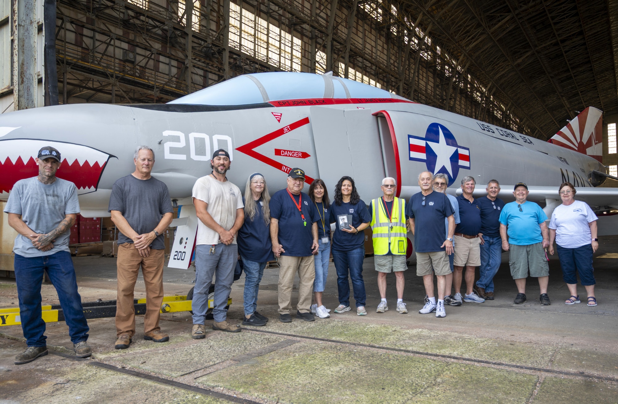 Volunteers who helped in the restoration and transport of the F-4 Phantom II stand in front of the historic military aircraft in Hangar 1 at Joint Base McGuire-Dix-Lakehurst, N.J., July 1, 2026. The restoration project, which took 13 months to complete, was accomplished by a team of 13 volunteers that included Airmen, Sailors, retired veterans, members of the Navy Lakehurst Historical Society and other civilians. (U.S. Air Force photo by Susan Moriarty) (This photo has been altered for security purposes by blurring out identification badges.)