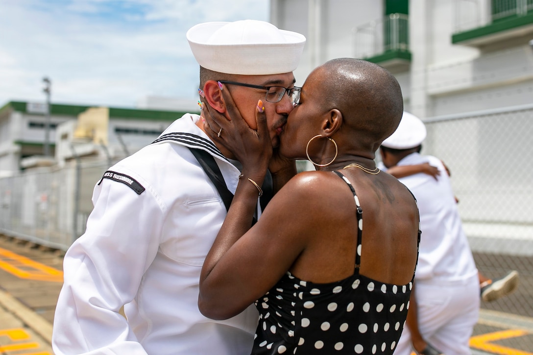 A sailor in a white uniform kisses a loved one in a black and white polka dot dress during the day.