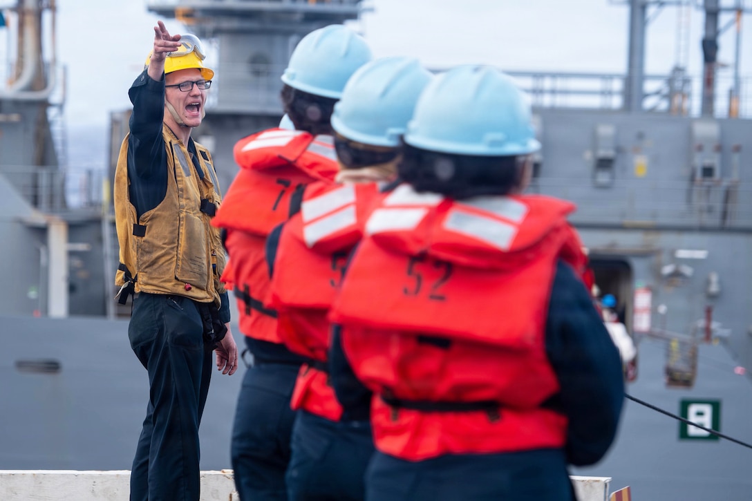 A sailor with a yellow hard hat and life vest shouts and points to sailors standing in formation wearing orange life vests with a ship in the background.