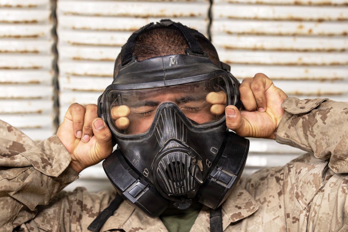 A close-up of a Marine Corps recruit in a camouflage uniform pulling off a black gas mask.