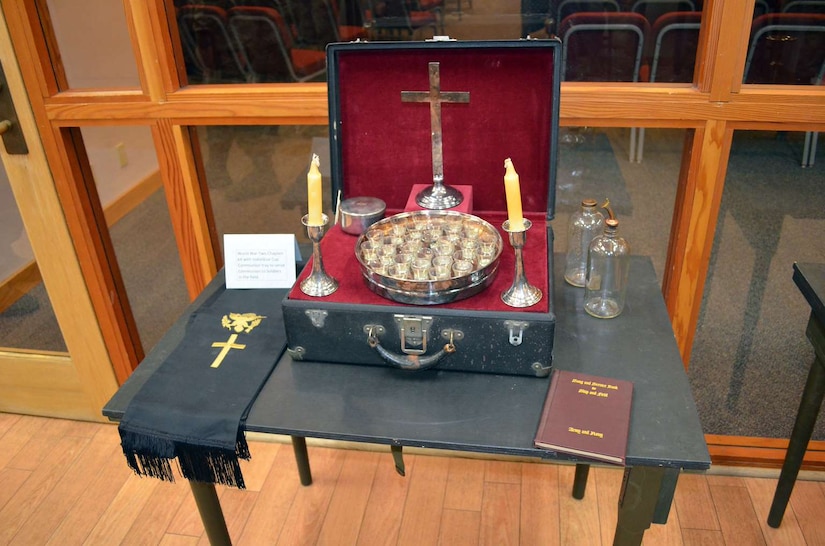 Candles, a cross and other religious materials are displayed on a table inside a room with wooden flooring.