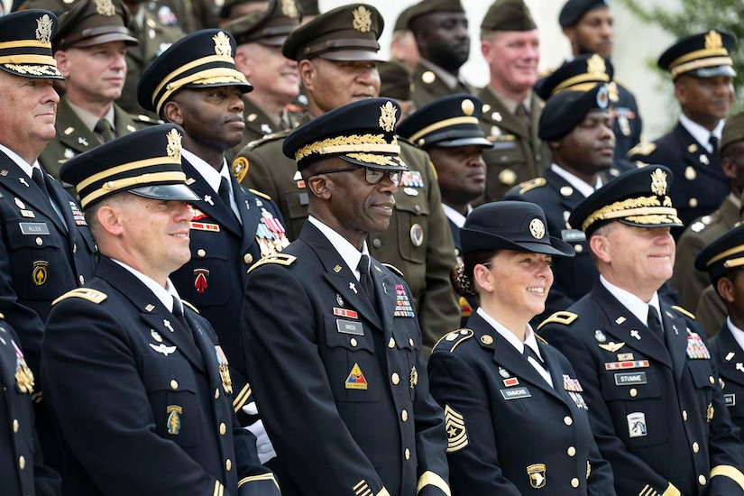 More than a dozen people in formal military uniforms stand in a group and pose for a picture outside.