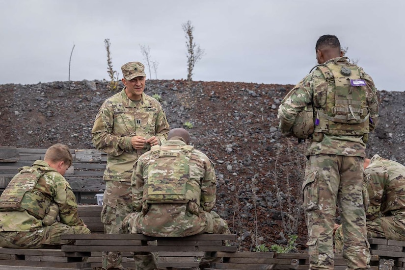 A man in a camouflage military uniform speaks to four other men in similar attire as they bow their heads outside.