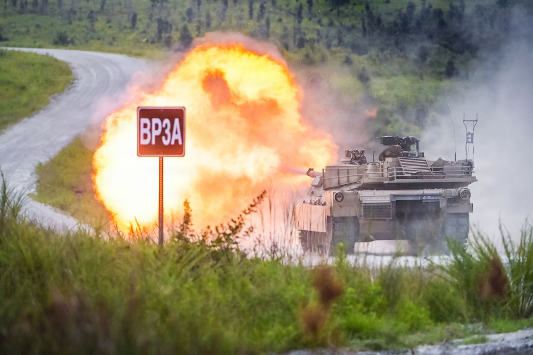 A tank with fire shooting out of it sits on a roadway at the bottom of a hill. Tall grass and a sign with BP3A on it are on the side of the road.