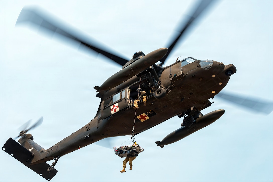 A person and rescue basket hang by a rope from a helicopter, and another person sits in the helicopter above holding the rope.